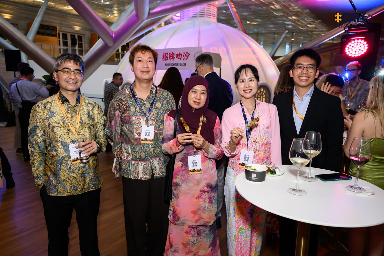 Five attendees, including Mr Goh Yihan SC, Deputy Attorney-General of Singapore, in traditional Southeast Asian attire pose together at the CALC Singapore 2026 Welcome Reception, with a signature laksa food station visible in the background.